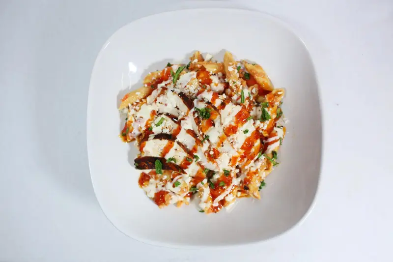 A dish of Baba’s Piri Piri Pasta Bake with chicken, topped with melted mozzarella, fresh spinach, and a garnish of herbs, served in a white bowl.