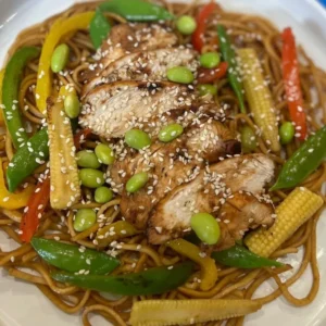 A bowl of Sticky Honey and Garlic Noodles topped with chicken, surrounded by colorful vegetables like peppers, baby corn, and sugar snaps.