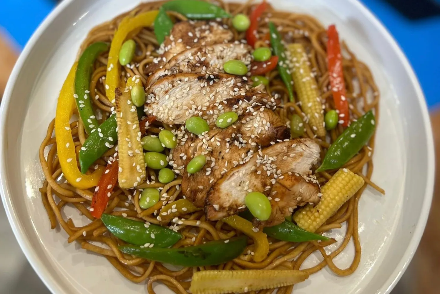 A bowl of Sticky Honey and Garlic Noodles topped with chicken, surrounded by colorful vegetables like peppers, baby corn, and sugar snaps.