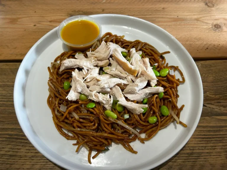 This dish is curry sauce chinese style noodles and features a serving of chinese style noodles, mixed with edamame beans and finely chopped onions, on a white round plate. Shredded pieces of tender chicken are layered on top, adding a hearty protein element. The meal is accompanied by a small cup of curry sauce, to enhance the flavour profile of the noodles and chicken. The presentation is simple and inviting, with a rustic, wholesome feel.