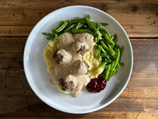 A plate of Swedish meatballs resting on a creamy bed of mash, generously smothered in a rich, velvety sauce. Served with a side of fresh green beans and a spoonful of tart cranberry sauce.