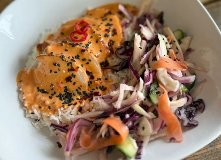 A white dish containing fluffy steamed rice topped with grilled, sliced chicken breast drizzled in a creamy orange Asian Bang Bang sauce and sprinkled with black sesame seeds, served alongside a crisp Asian salad of shredded cabbage, carrot ribbons, red onion and cucumber.