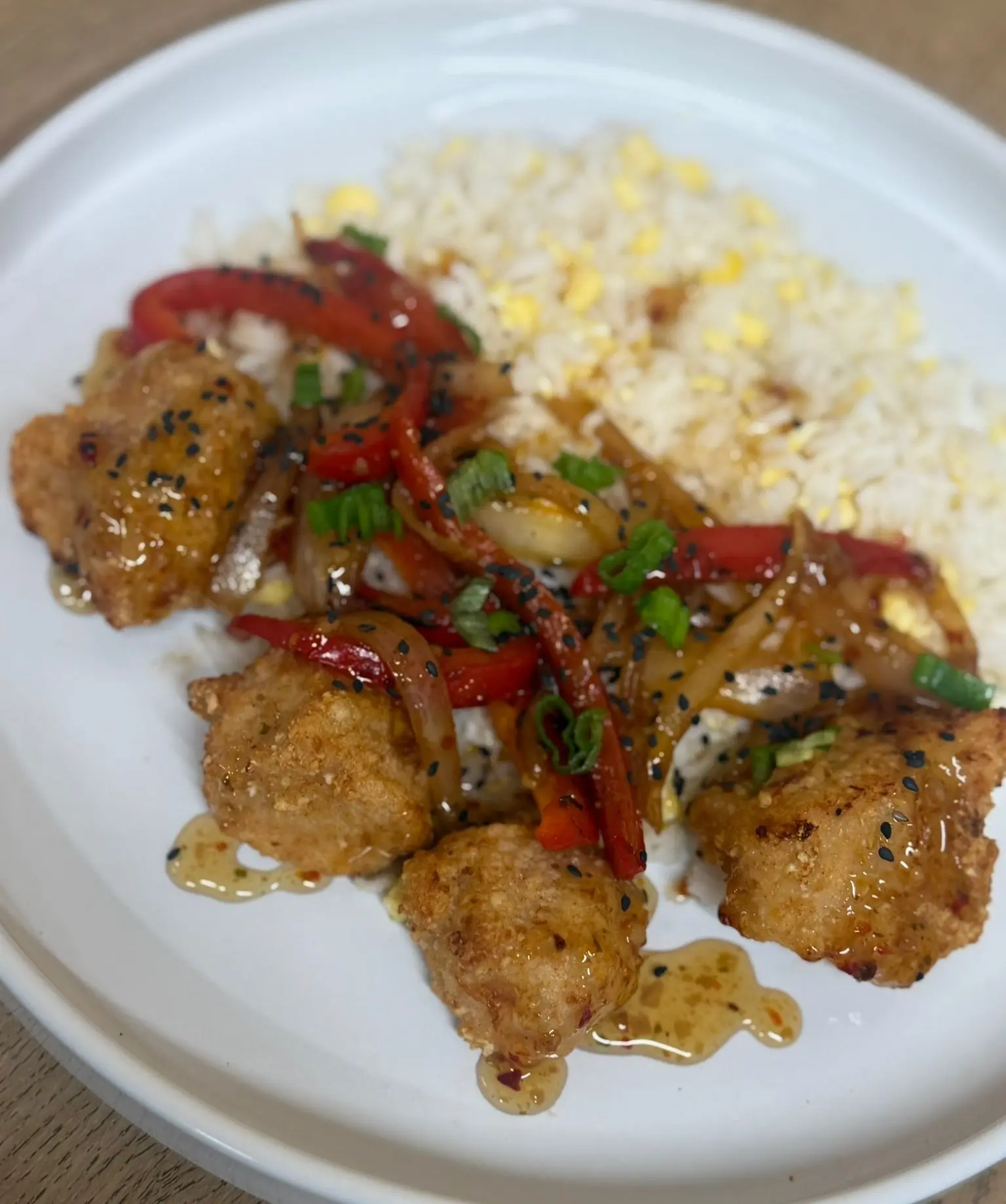 Crispy honey-glazed chicken bites served with egg-fried rice, sautéed red peppers and onions, topped with spring onions and black sesame seeds on a white plate, by Savage Kitchen